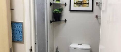 Une petite salle de bain avec des toilettes, une douche et un lavabo. Les murs sont décorés de carreaux et un tableau encadré est accroché au-dessus des toilettes.