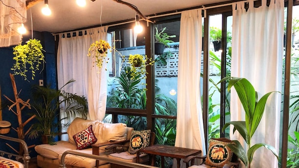 A cozy room with a bed, sofa, and chairs, featuring a large window with curtains and various potted plants. - Nego Home Ancient Hotel (Bangkok)