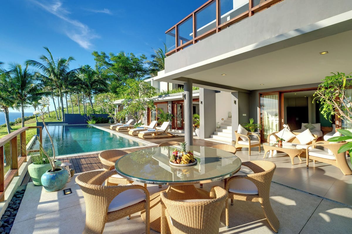 Un área de piscina exterior moderna con sillas de descanso y una mesa de vidrio, rodeada de plantas tropicales y vista al océano.