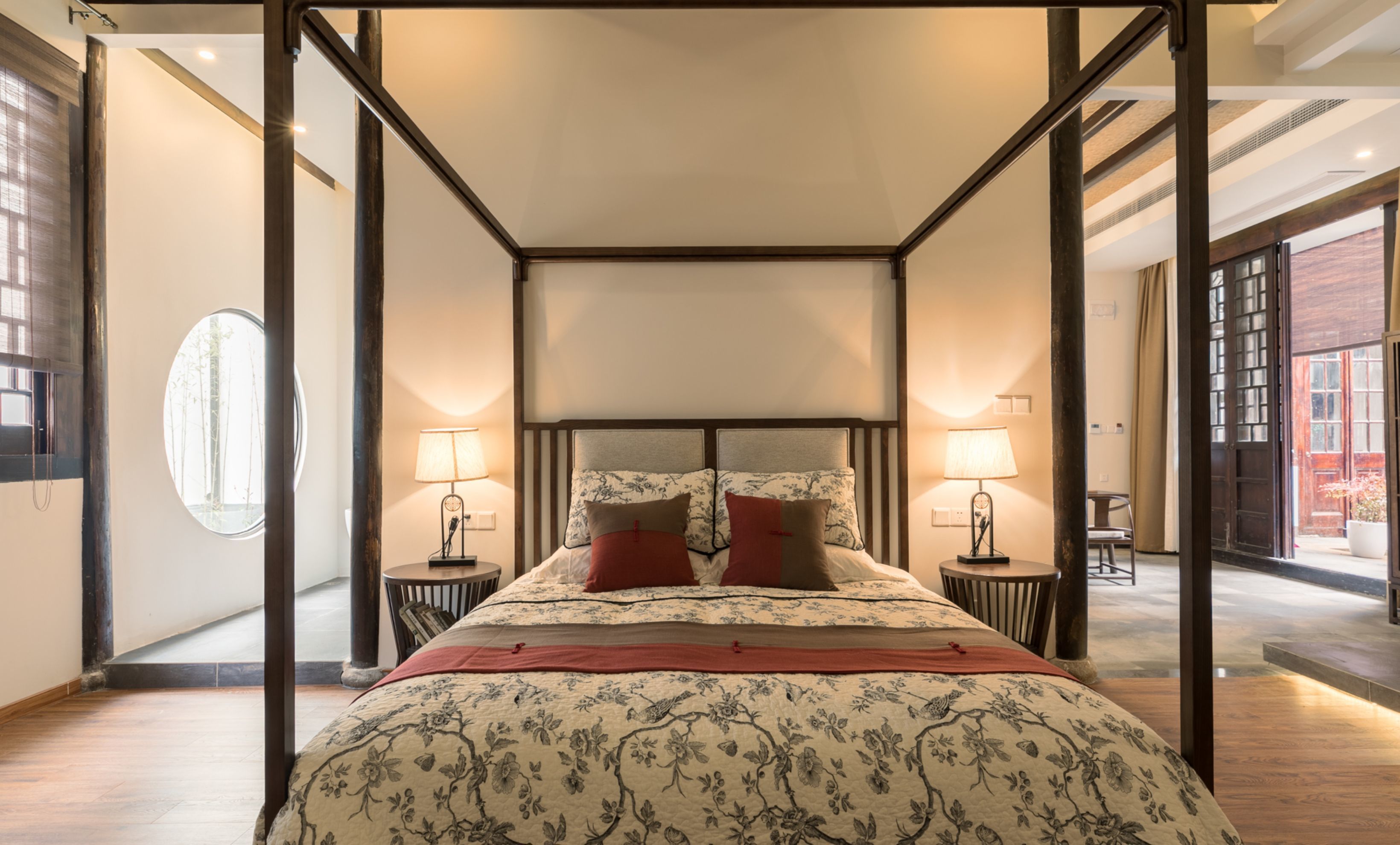 A four-poster canopy bed with floral bedding, two bedside tables with lamps, and a view of a brick building through an open door.
