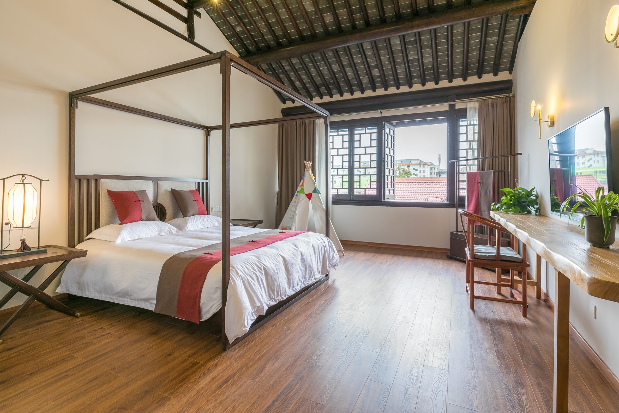 A four-poster bed with a wooden frame, a desk with a chair, and a view of buildings through a window.