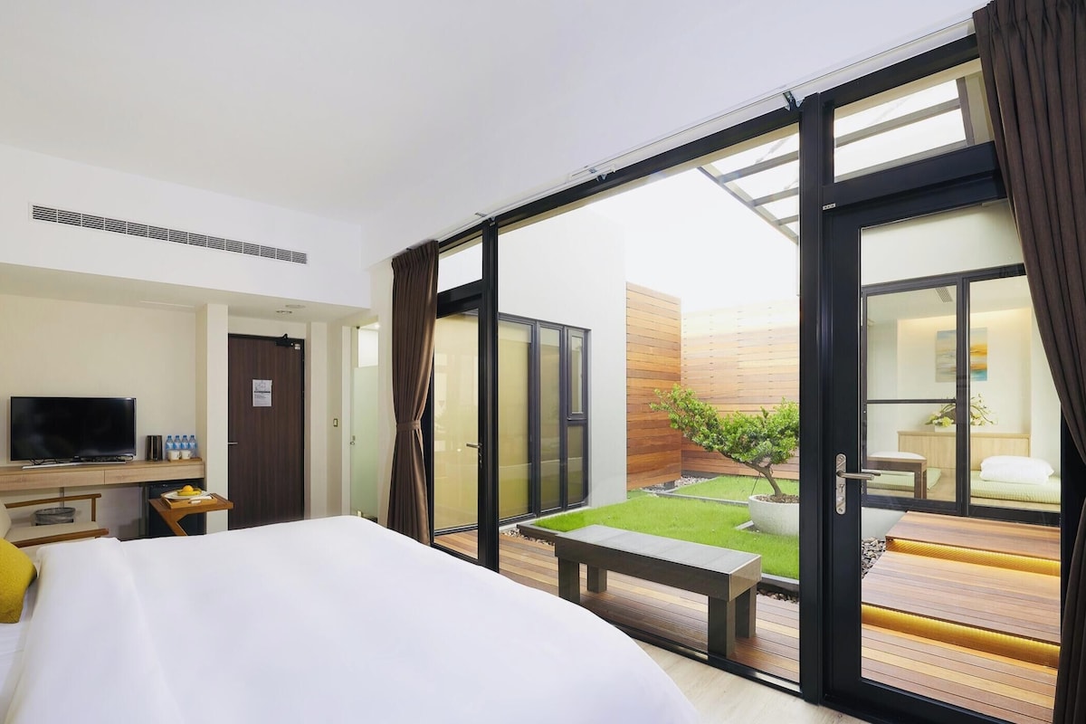 A hotel room with a bed, a desk with a TV, a small table with a tray of fruit, and a balcony with a view of a garden.