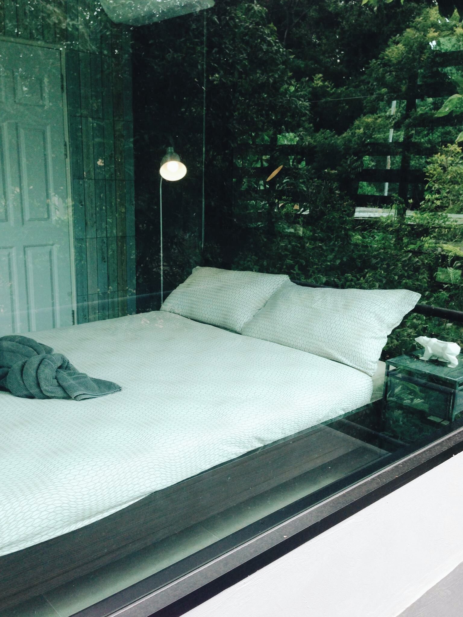 A bed with a white mattress and pillows, a bedside table with a white figurine, and a window with a view of greenery.