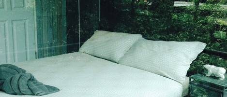 A bed with a white mattress and pillows, a bedside table with a white figurine, and a window with a view of greenery.