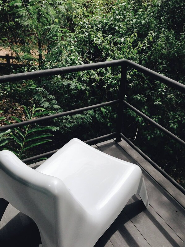 A white plastic chair on a wooden deck with a metal railing, surrounded by dense green foliage.