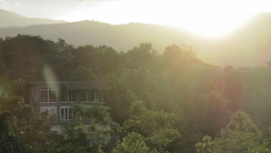 Una casa de dos pisos rodeada de un bosque frondoso, con una cadena montañosa al fondo.