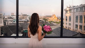 Une personne tenant un bouquet de fleurs, regardant par une fenêtre donnant sur un paysage urbain avec des immeubles et un temple.