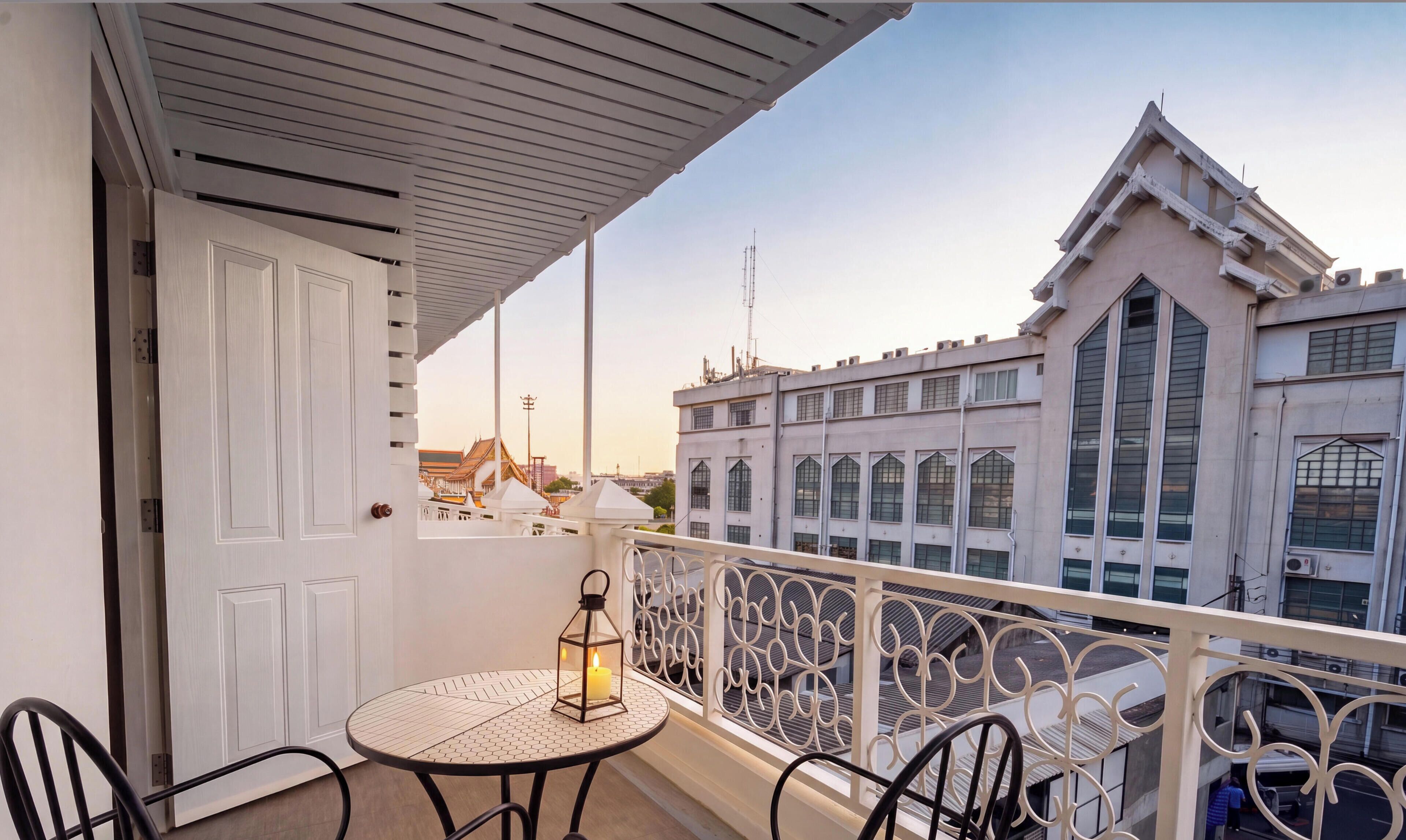 Uma varanda com mesa e cadeiras, uma porta branca e vista para um edifício com grandes janelas.