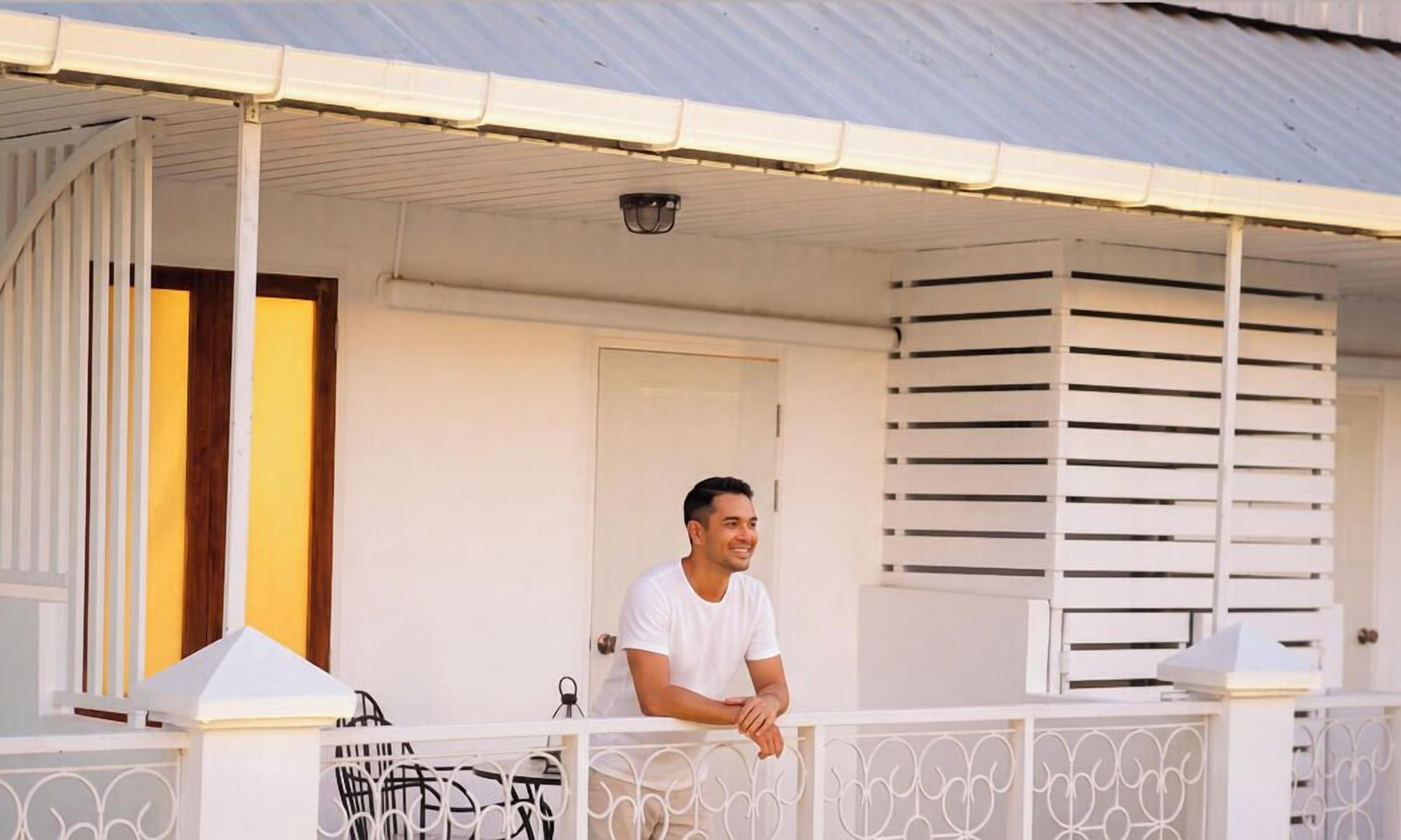 A man sitting on a balcony with a white railing, a door, and a window with blinds.