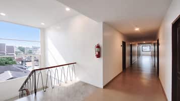 Un pasillo de hotel con un extintor en la pared, una barandilla y vistas de la ciudad a través de amplios ventanales.