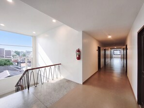 A hotel corridor with a fire extinguisher mounted on the wall, a handrail, and a view of the city through large windows.