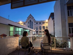 Two individuals seated on a balcony with a view of a building with large windows and a distinct architectural style.