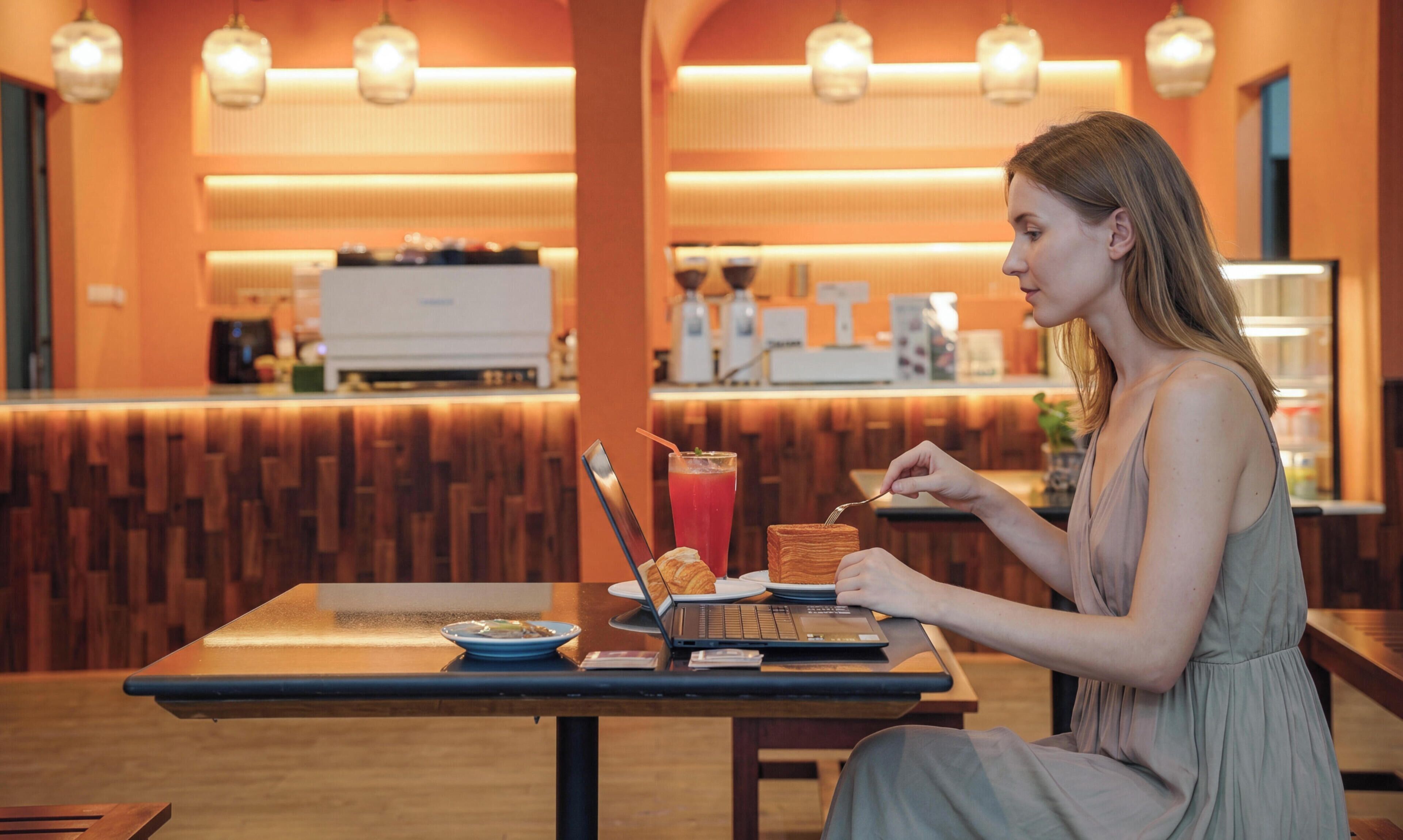 Una mujer sentada a una mesa con una computadora portátil, un vaso de bebida roja y un plato con comida y un croissant.
