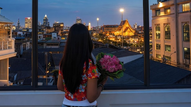 Une personne tenant un bouquet de fleurs, regardant par une fenêtre donnant sur un paysage urbain avec des bâtiments et une imposante structure dorée.