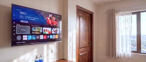 Quarto de hotel com televisão de tela plana na parede, escrivaninha de madeira com bandeja contendo garrafas de água e um controle remoto, uma porta e uma janela com cortinas.