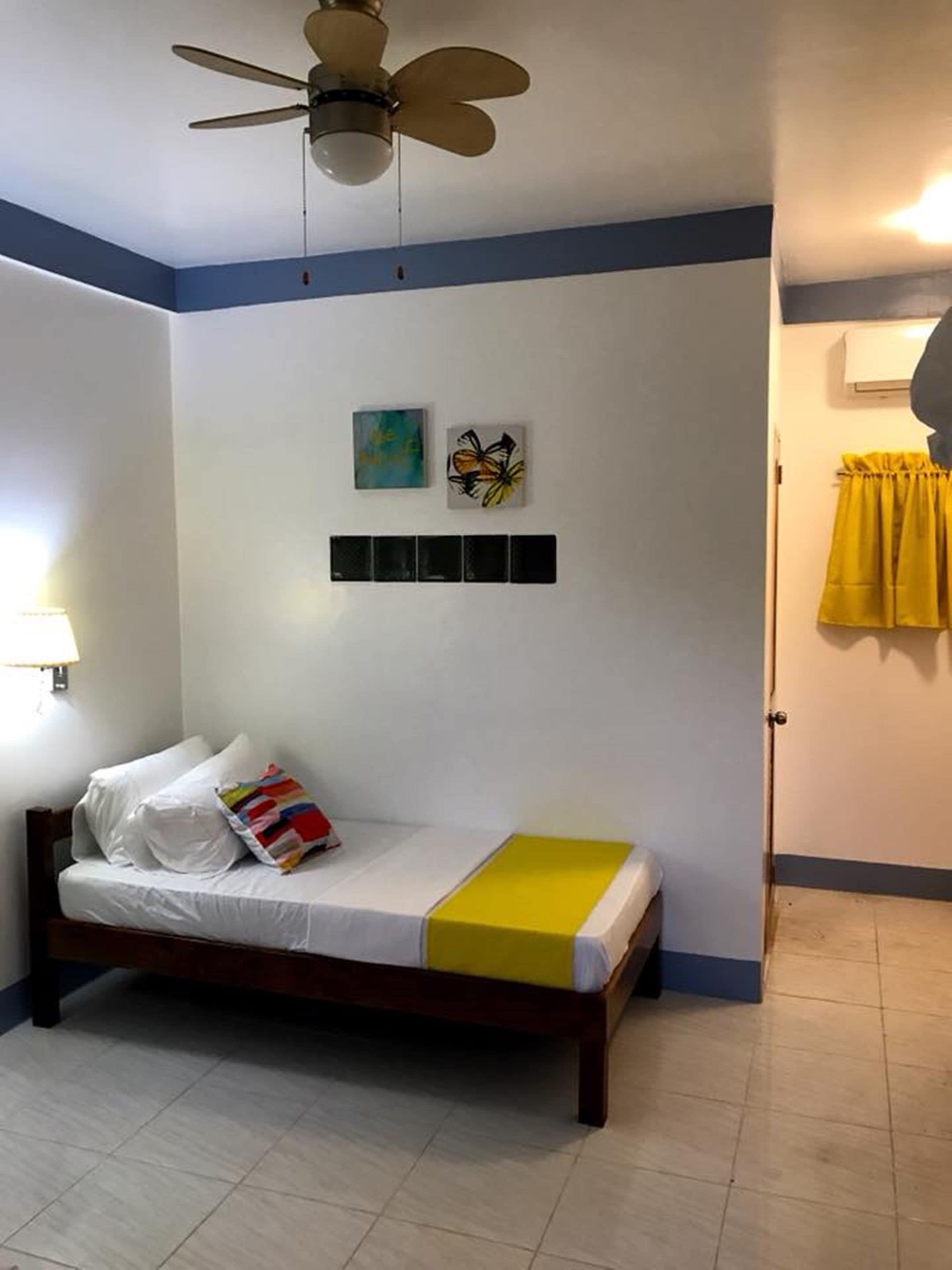 Un dormitorio con cama, ventilador de techo, aire acondicionado instalado en la pared y una puerta con cortinas amarillas.