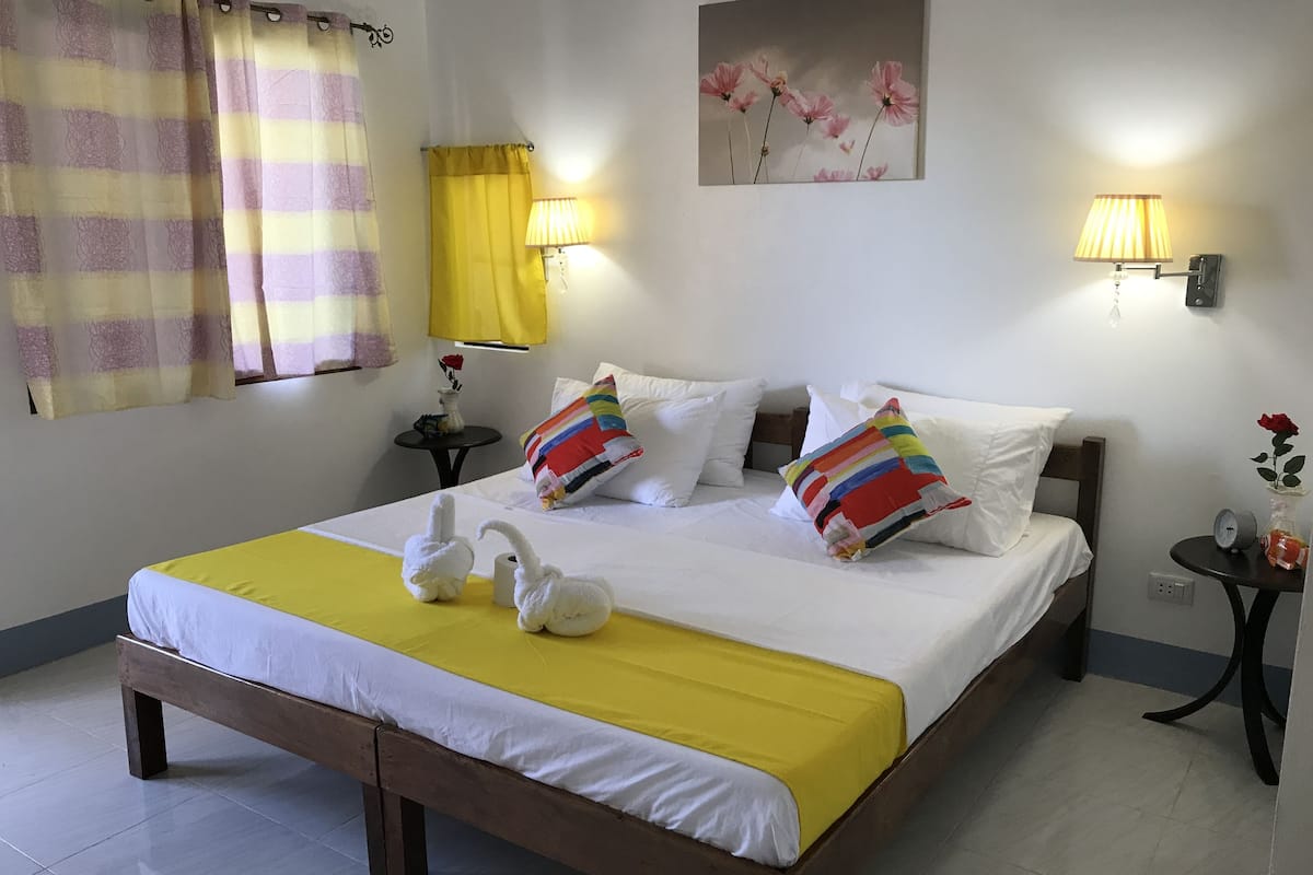 A neatly made bed with white bedding and a yellow accent, a small bedside table with a lamp, and a wall-mounted picture of flowers.