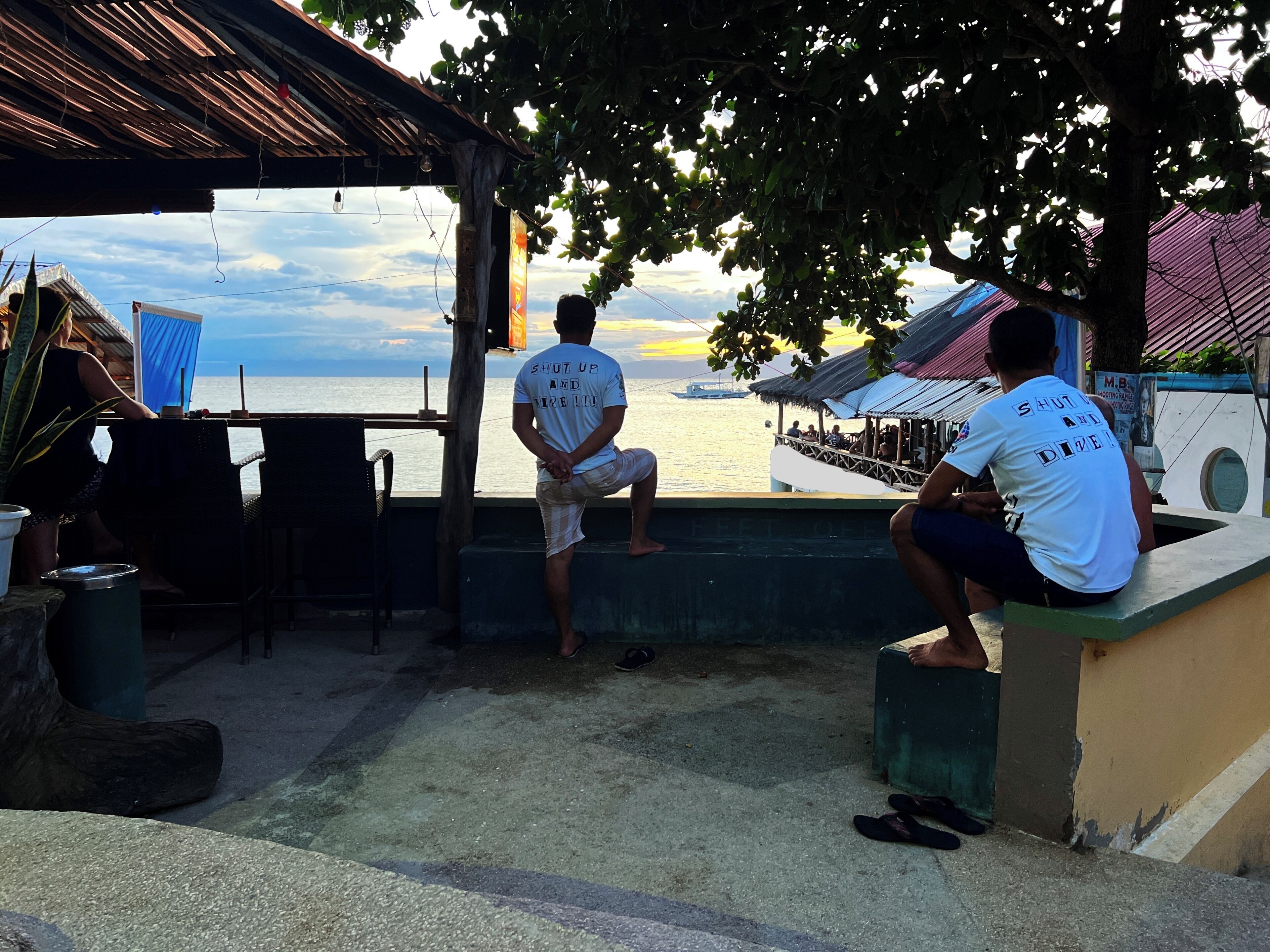 A coastal outdoor seating area with a view of the sea, featuring a wooden pergola and a thatched roof structure.