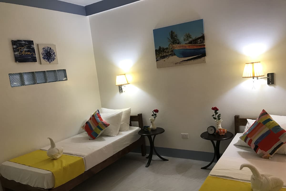 A hotel room with two single beds, each with a colorful pillow and a swan-shaped pillow. There are two small tables with lamps, a painting of a beach scene, and a wall-mounted light fixture.