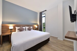 A modern hotel room with a large bed, wooden headboard, bedside lamps, a flat-screen TV, and a view of greenery through the window.