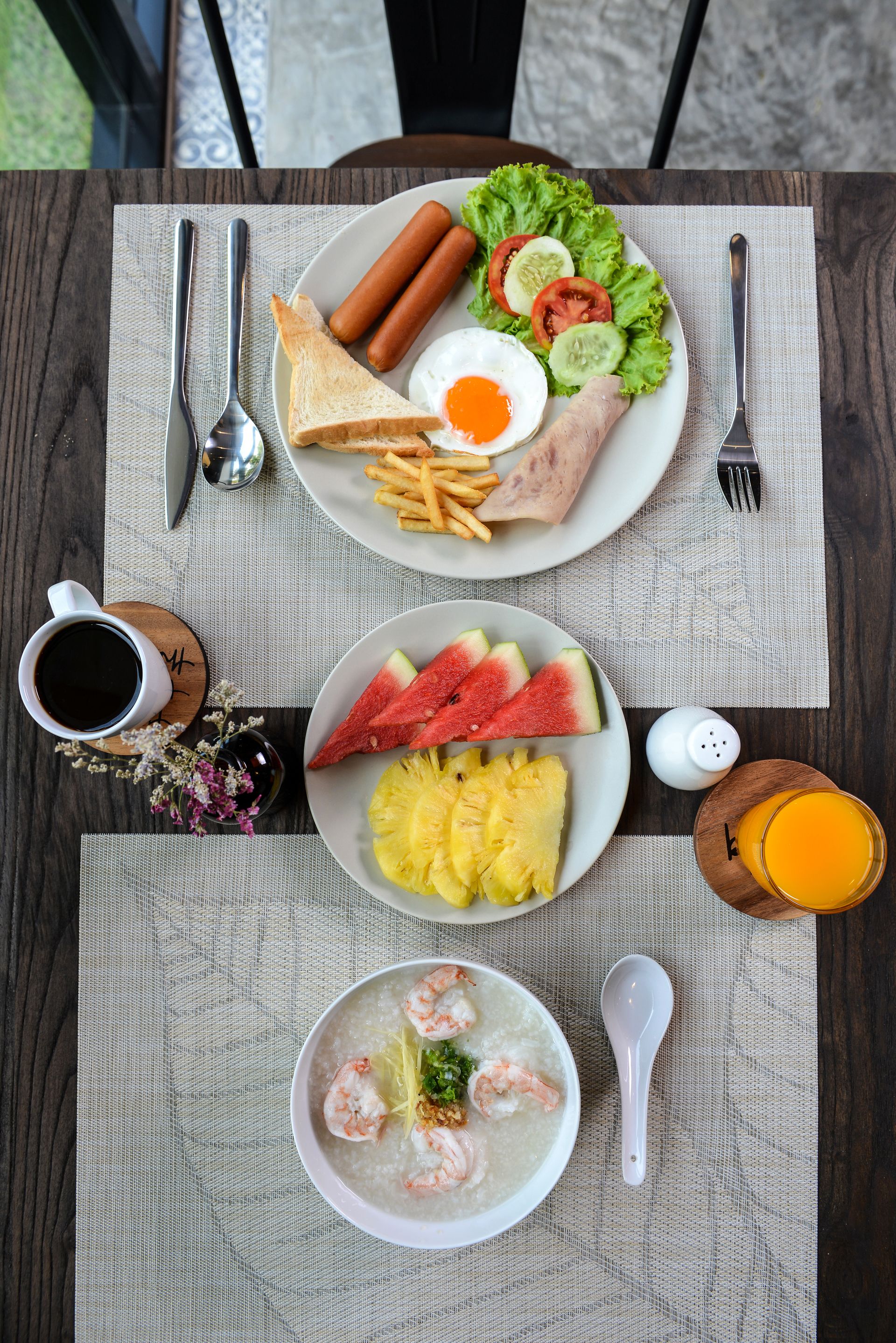 A table with a plate of breakfast including toast, sausages, eggs, and salad; another plate with fruit and a bowl of soup; and a cup of coffee.