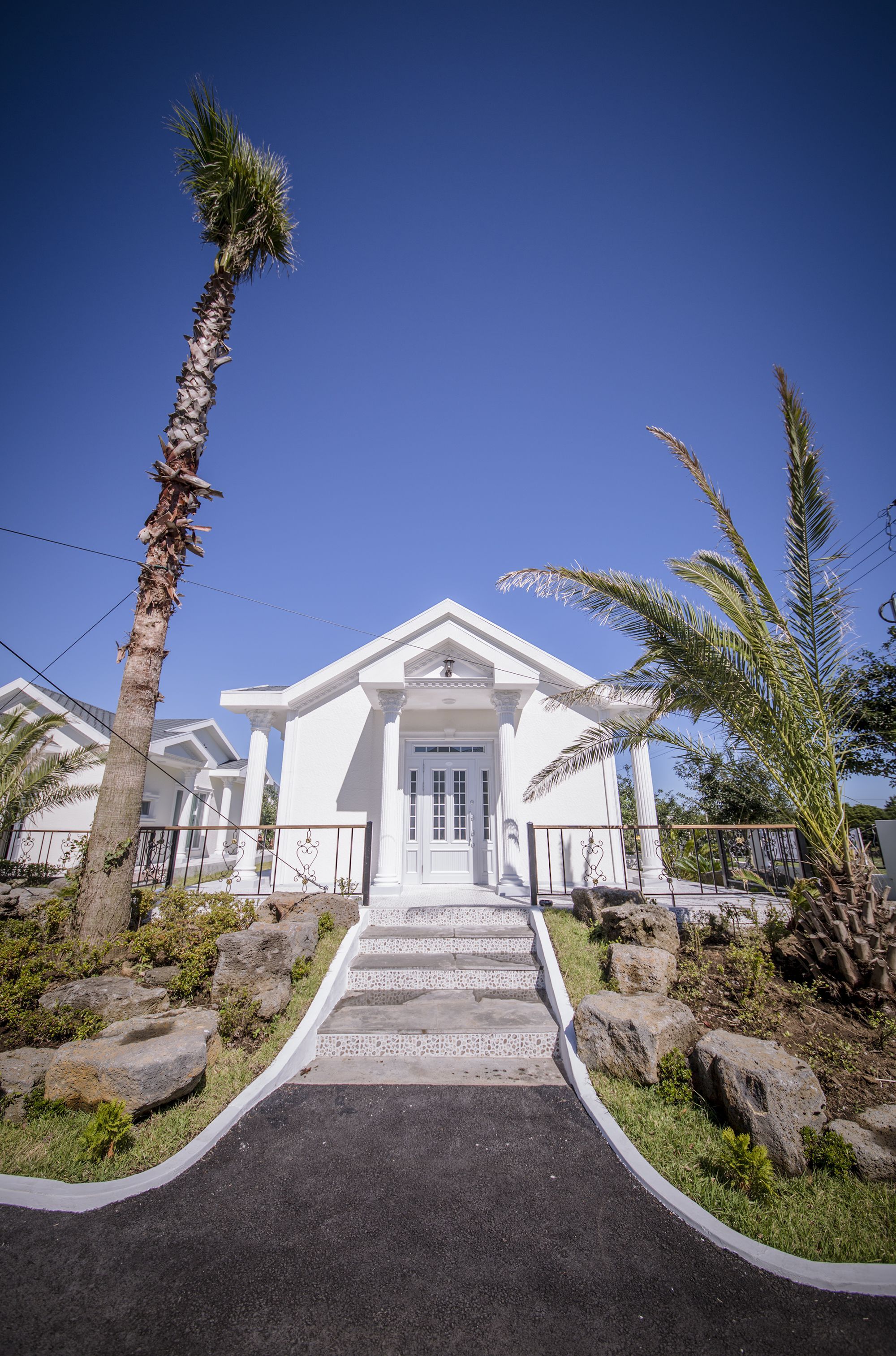 A white building with a central entrance, flanked by palm trees and surrounded by rocks.