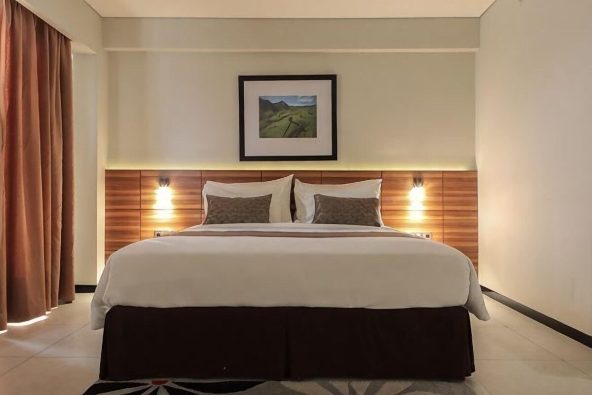 Habitación de hotel con una cama grande, cabecera con iluminación integrada, un cuadro enmarcado en la pared y una ventana con cortinas.