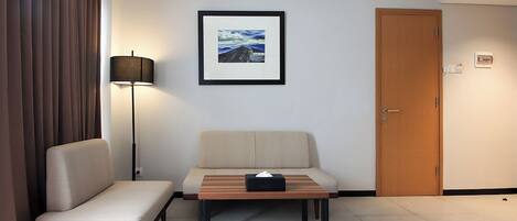 A hotel room with a sofa, a small table with a tissue box, a framed picture on the wall, a door, and an air conditioning unit.