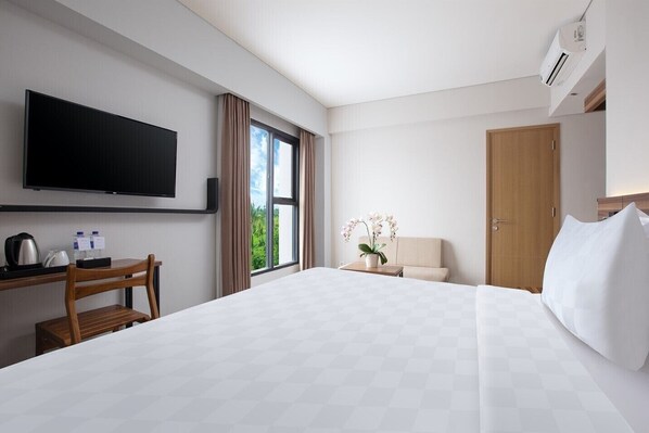 A hotel room with a bed, a desk with a chair, a television, a window with curtains, a potted plant, and a door.