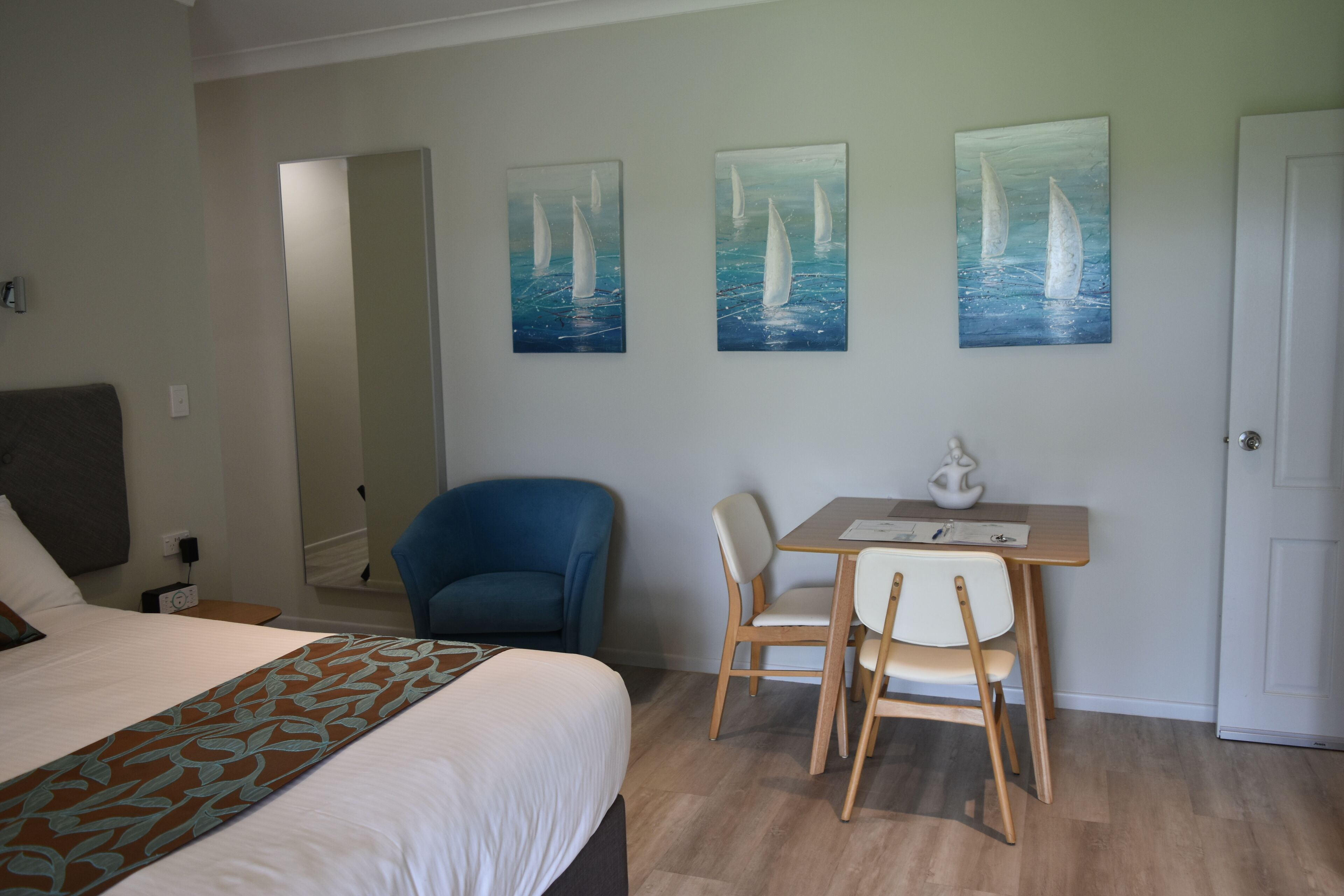 A hotel room with a bed, a desk with two chairs, and three paintings of sailboats on the wall.