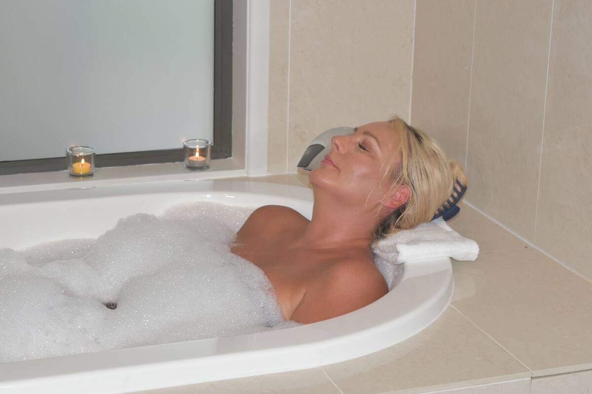 A person relaxing in a bathtub filled with bubbles, with a towel and a hairdryer on the side.
