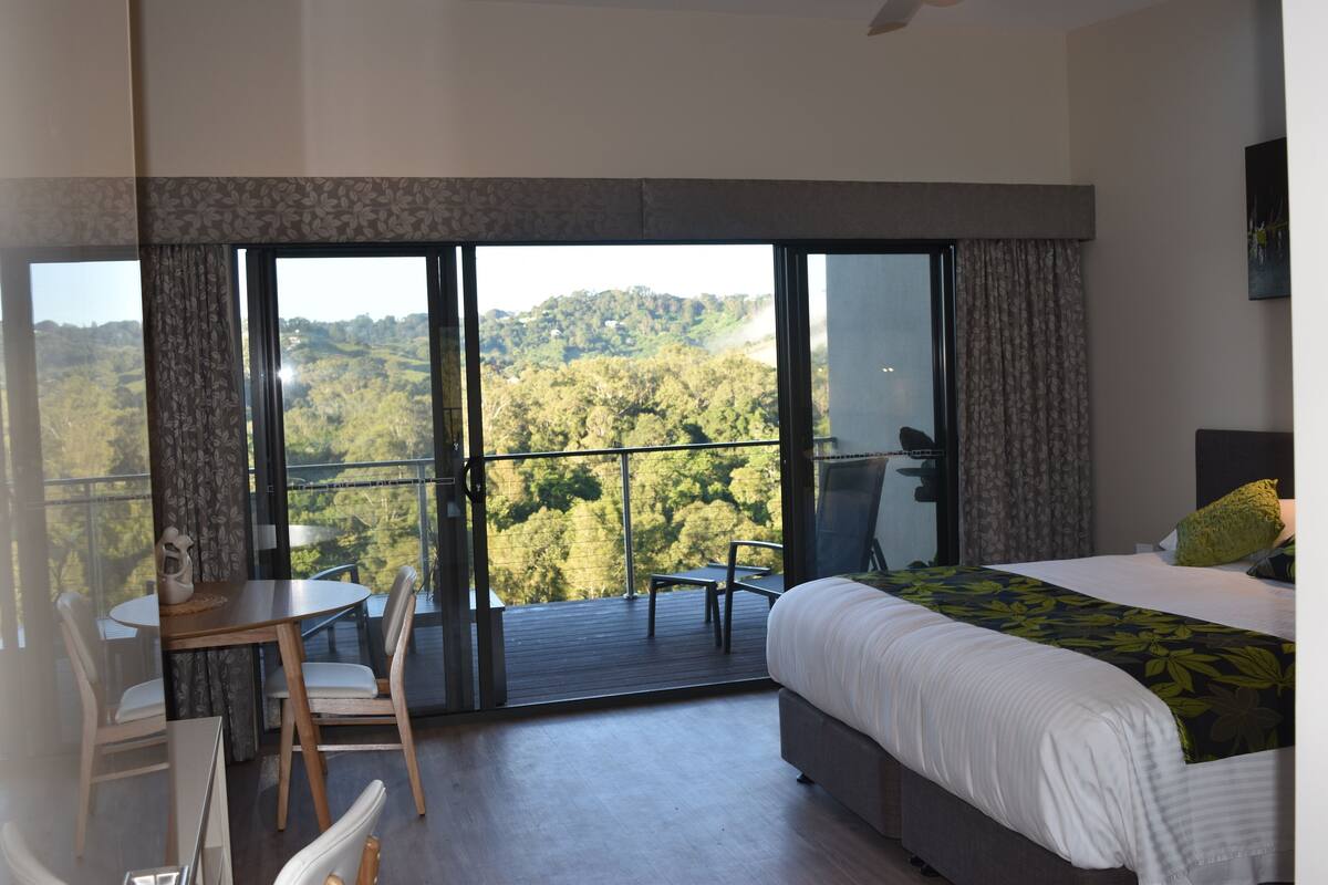 A bedroom with a large bed, a desk, and a chair. There is a balcony with a view of a forested area.
