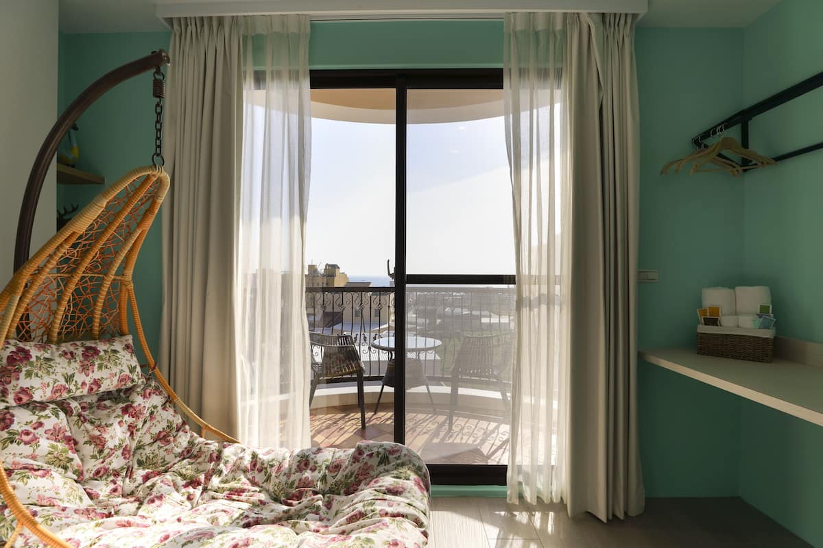 Quarto com cama, cadeira suspensa, varanda com mesa e cadeiras, e vista para o exterior.