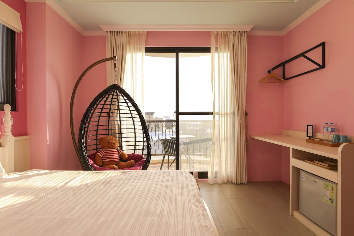 Quarto com cama, uma cadeira suspensa com um urso de pelúcia, uma mesa pequena e vista para o exterior.