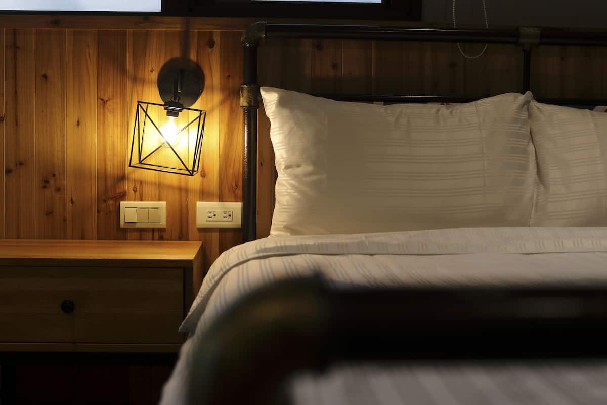 Quarto com painéis de madeira, uma cama, um criado-mudo e um aplique na parede.