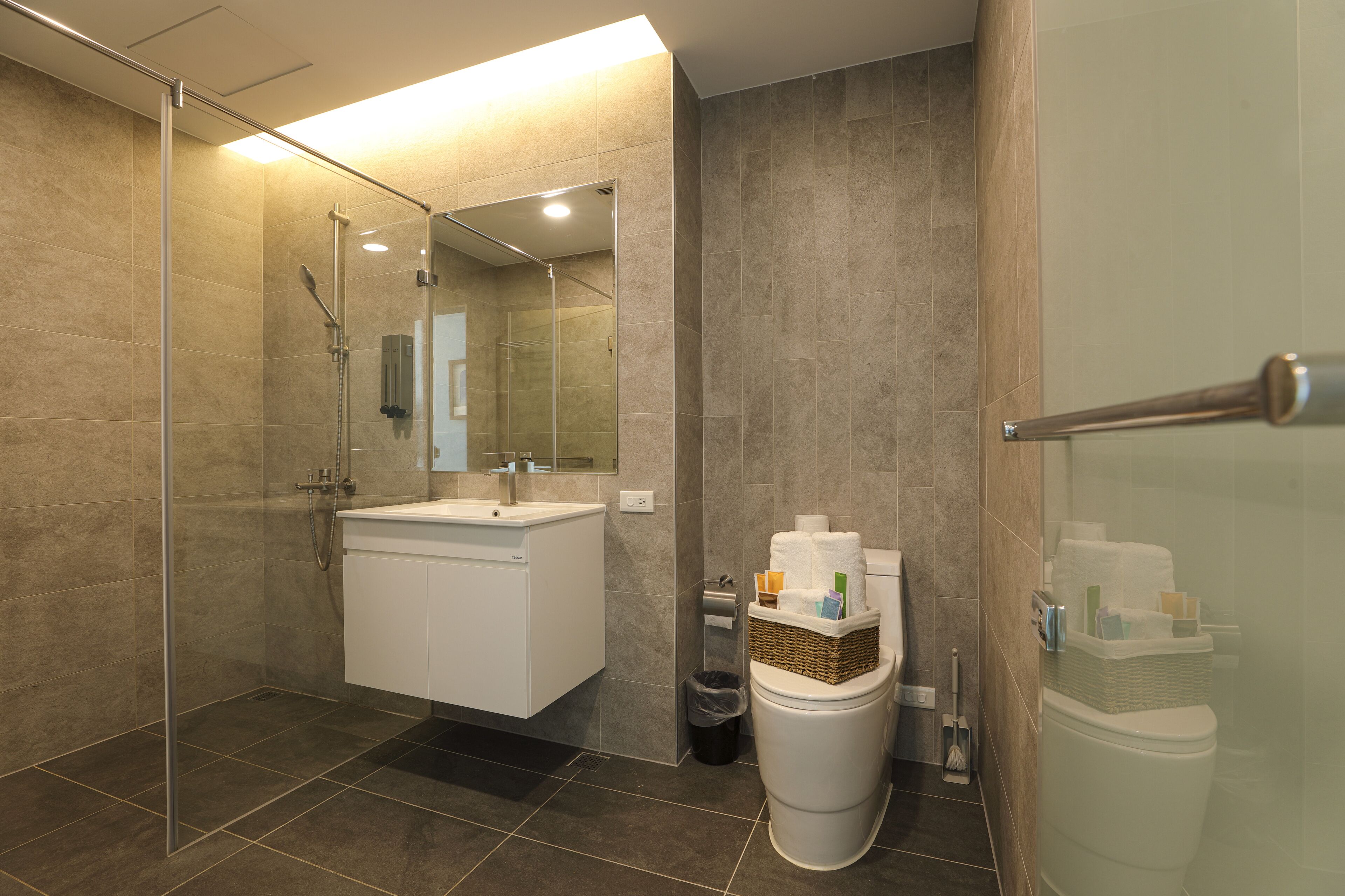 Un baño moderno con cabina de ducha de vidrio, un lavabo blanco y un inodoro con una cesta de artículos de aseo.