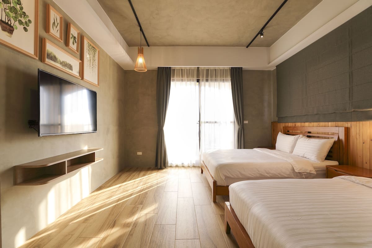 Quarto moderno com duas camas, uma televisão de tela plana, piso de madeira e uma janela grande com cortinas.