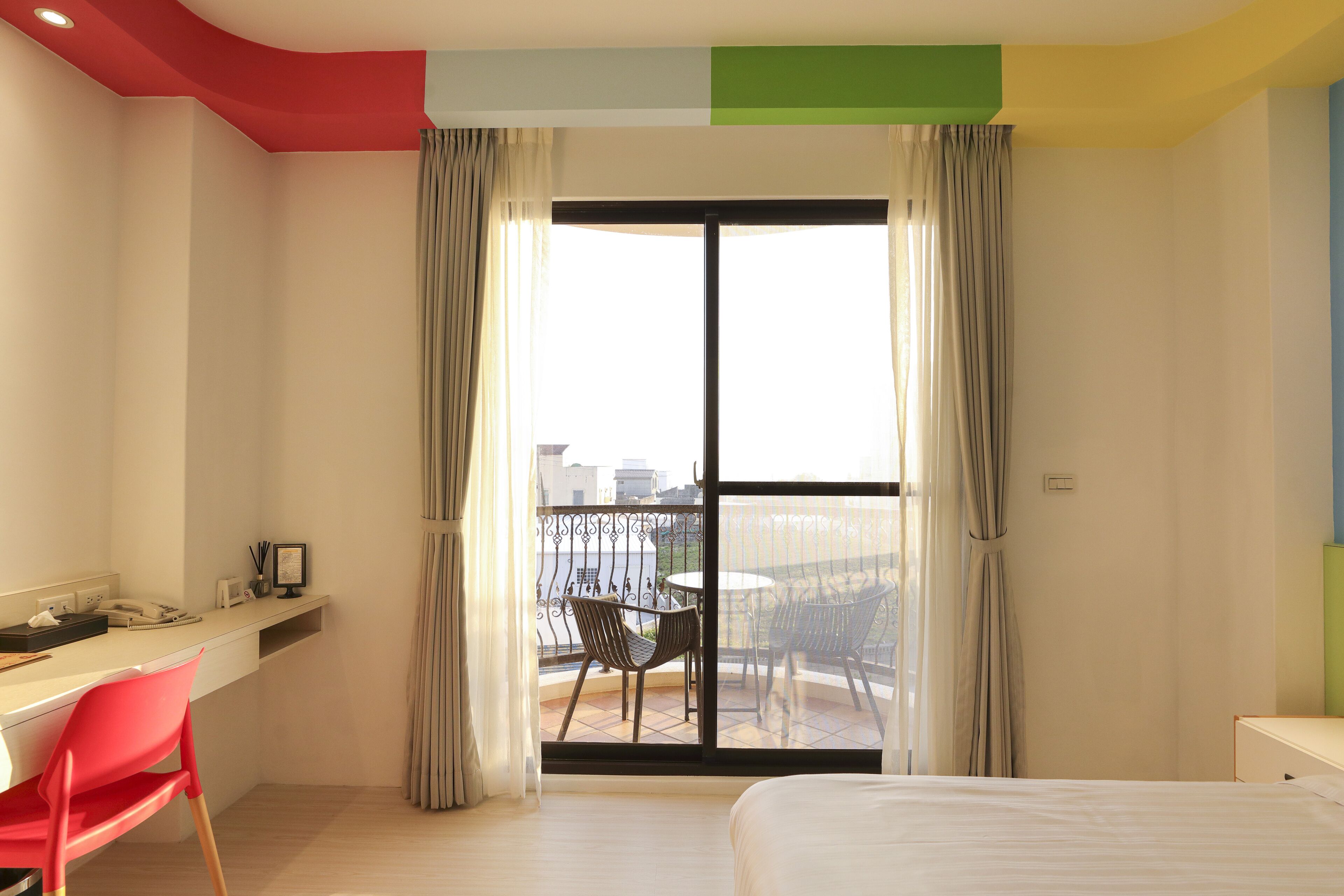 A hotel room with a bed, a desk with a chair, a balcony with a table and chairs, and a view of the city.