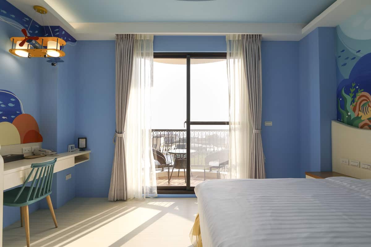 Quarto de hotel com cama, secretária com cadeira, varanda com área de estar e uma janela grande com cortinas.