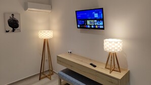 A modern room with a wooden console table, two floor lamps, a mounted TV displaying a home screen, and a wall-mounted air conditioner. - Aphrodite Studios (Kalymnos)