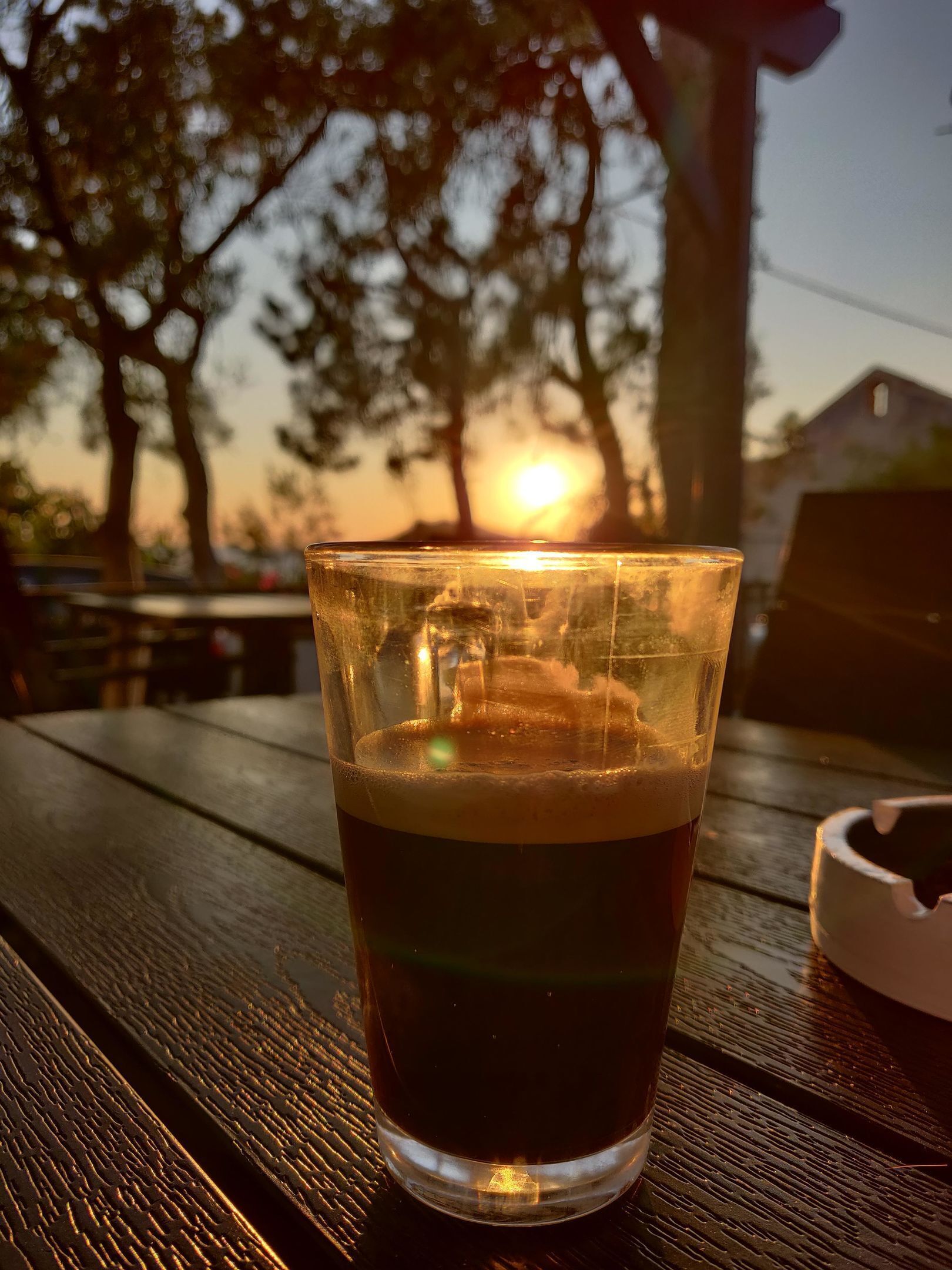 Un vaso de líquido oscuro sobre una mesa de madera, con un atardecer de fondo.