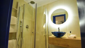 A glass-enclosed shower with a handheld showerhead, a wooden vanity with a blue bowl sink, and a round mirror with a blue frame. - Aphrodite Studios (Kalymnos)