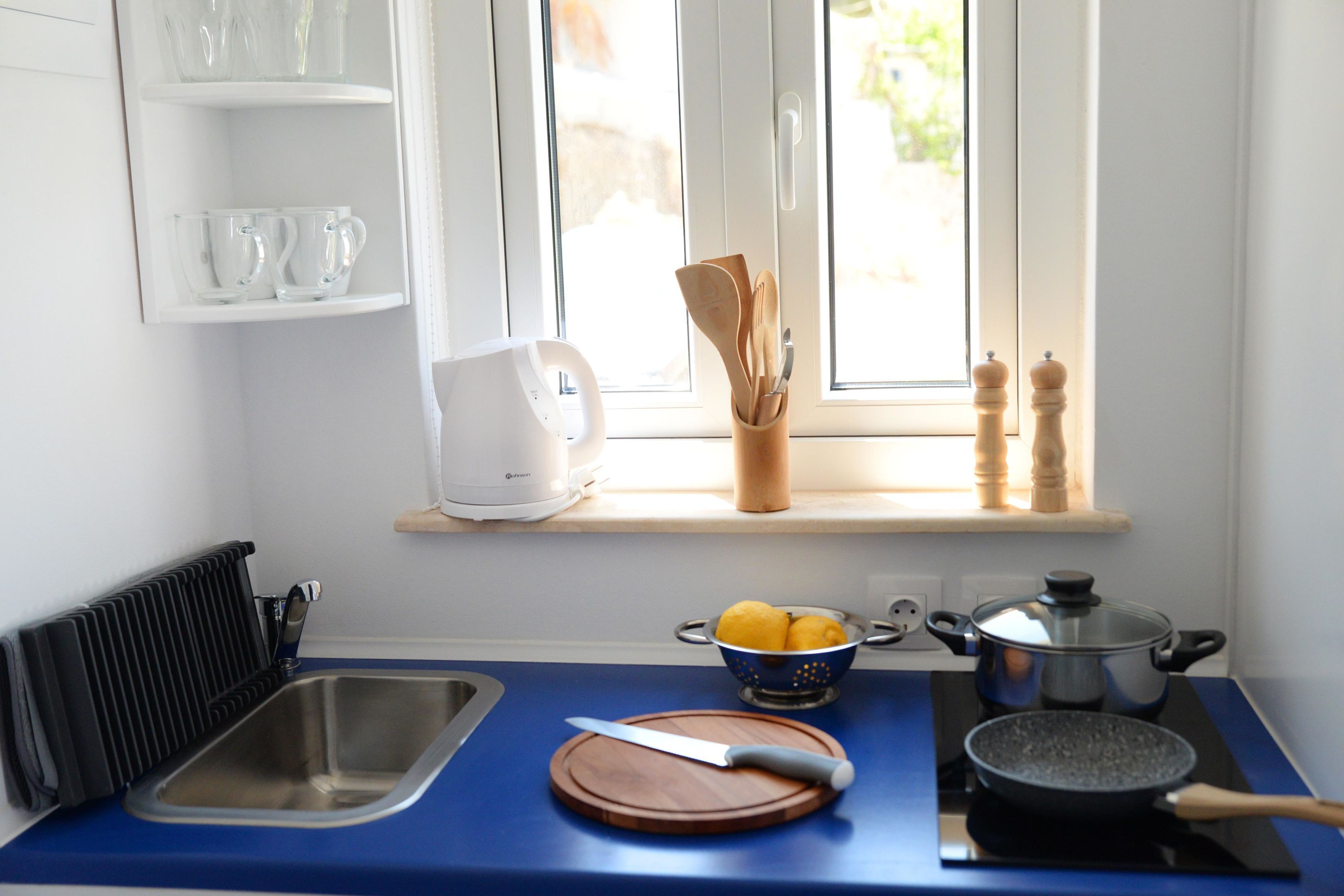 A kitchen with a blue countertop, a sink, a cutting board with a knife, a pot on an induction cooktop, and a window with a view of greenery.