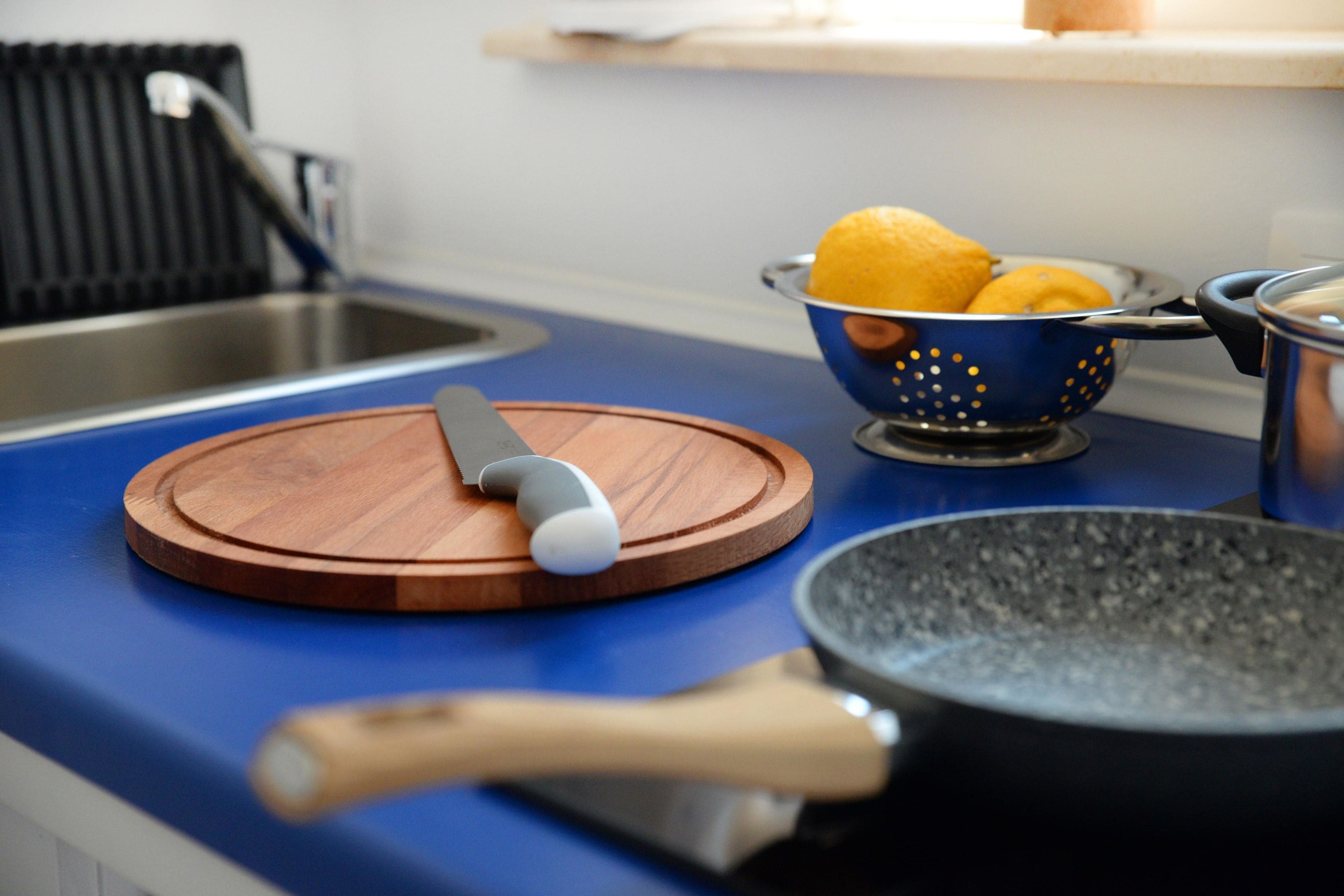 A kitchen counter with a cutting board, a peeler, a colander with lemons, and a frying pan.