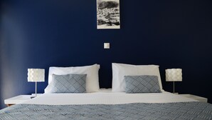 A neatly made double bed with white and patterned pillows, a blue patterned bedspread, and two bedside lamps with white shades. - Aphrodite Studios (Kalymnos)