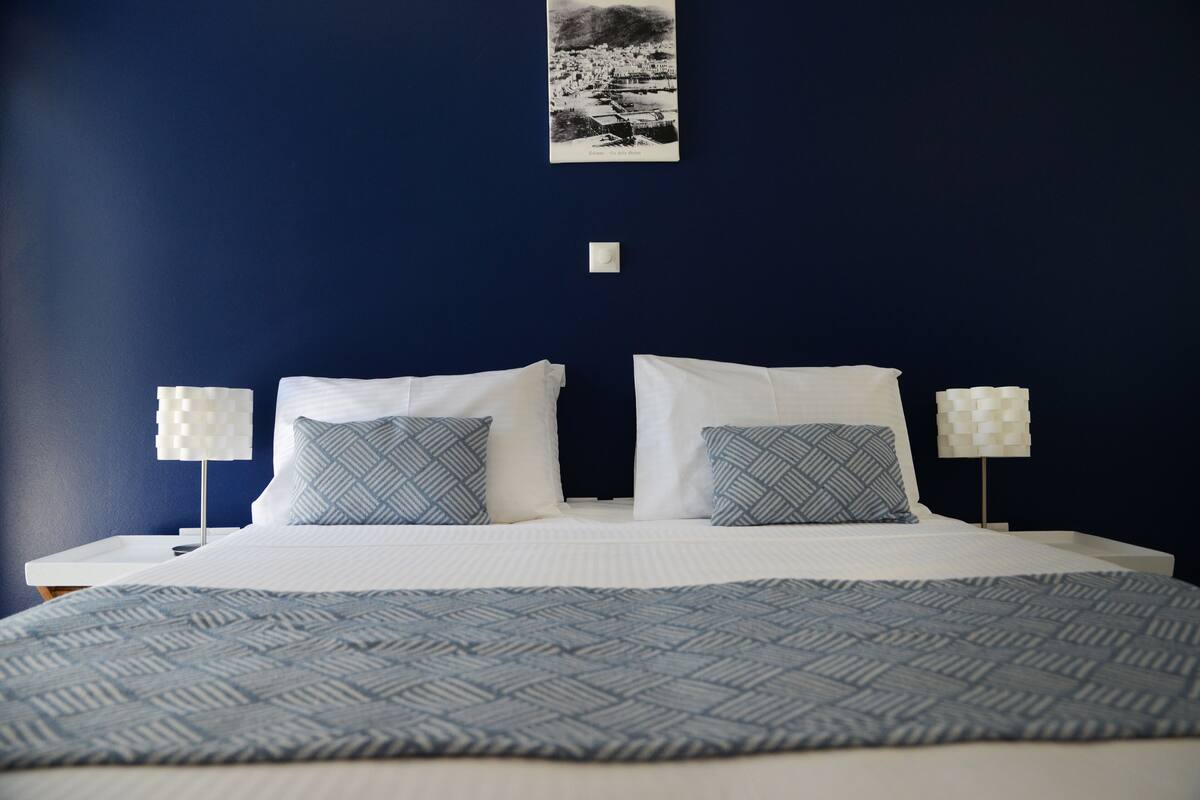 A neatly made double bed with white and patterned pillows, a blue patterned bedspread, and two bedside lamps with white shades.