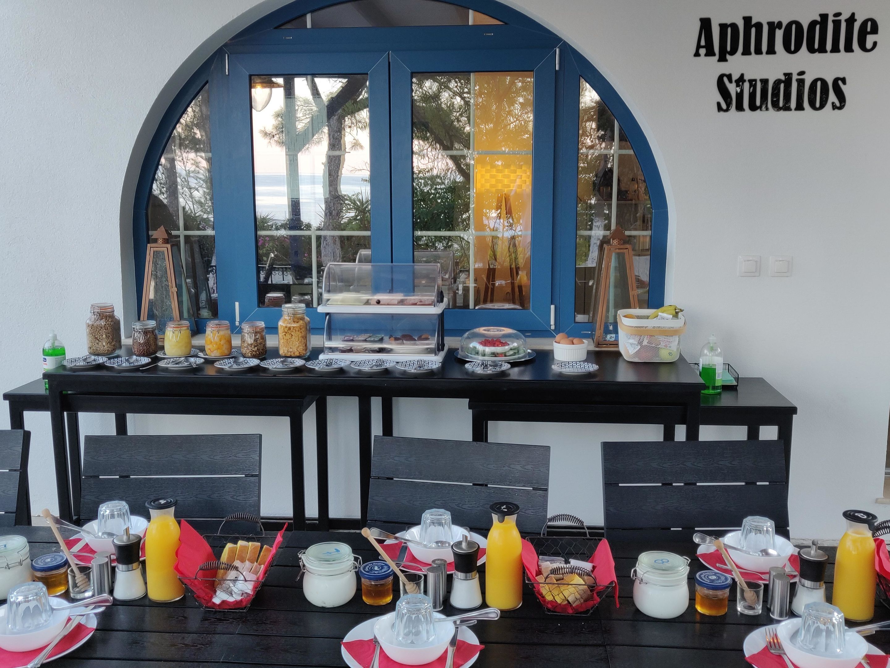 Ein Frühstückstisch mit verschiedenen Speisen und Beilagen, ein Fenster mit Blick und der Schriftzug 'Aphrodite Studios' an der Wand.