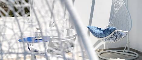 A white hanging chair with a blue cushion, a clear glass table, and a white chair with a blue cushion.