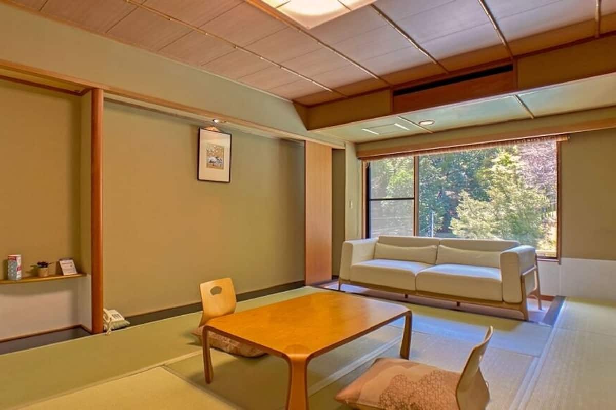 Habitació d'estil japonès tradicional amb fons de tatami, taula de fusta baixa, sofà i finestra amb vista a arbres.