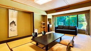 Ein traditioneller japanischer Raum mit Tatami-Böden, einem niedrigen Tisch, einer Couch und einem großen Fenster mit Blick auf Grünflächen.